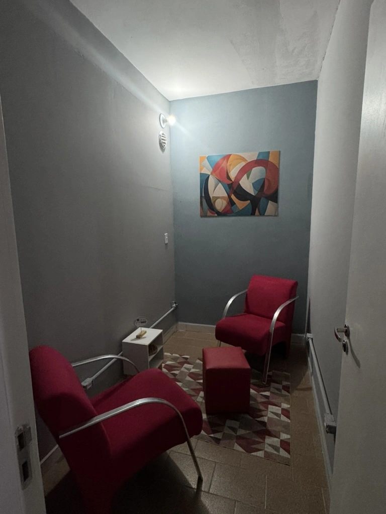 Sala de Psicologia com duas poltronas vermelhas, mesa lateral, tapete geométrico e quadro colorido.