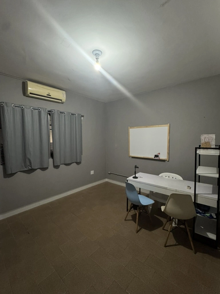 Segundo ângulo da Sala 2 - Auditório, com mesa, quadro branco, cadeiras e estante.